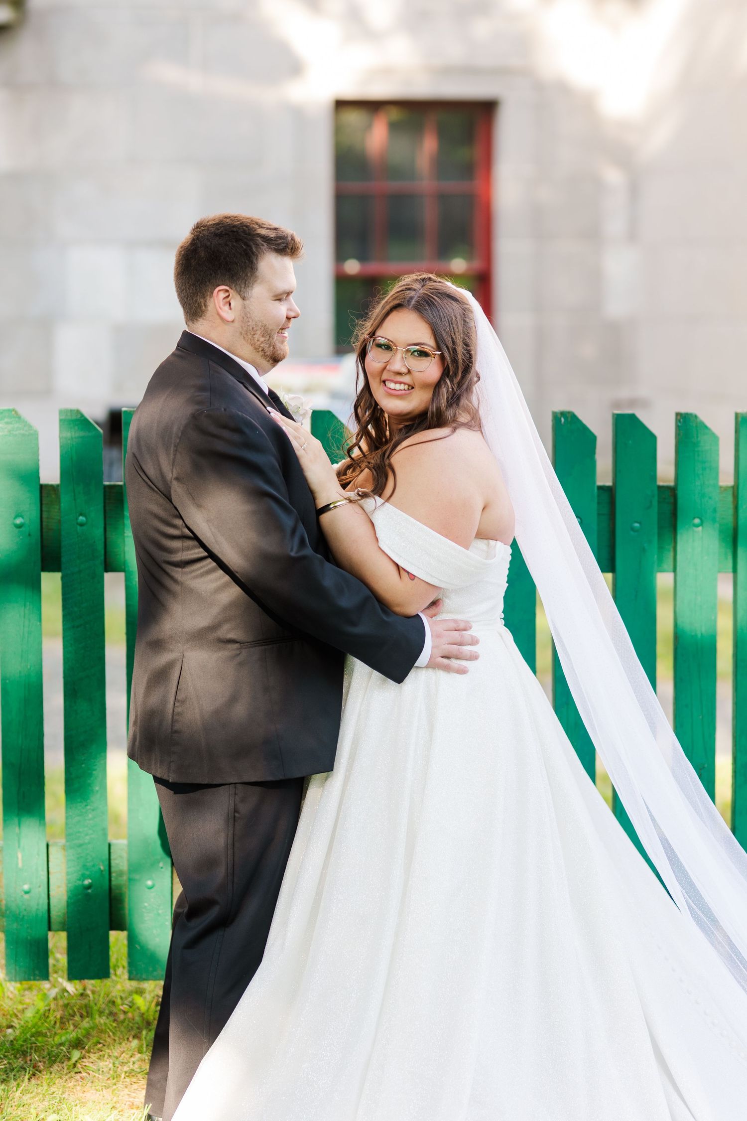 A bride and groom share intimate romantic moments in front of a green picket fence during their wedding photo session.
