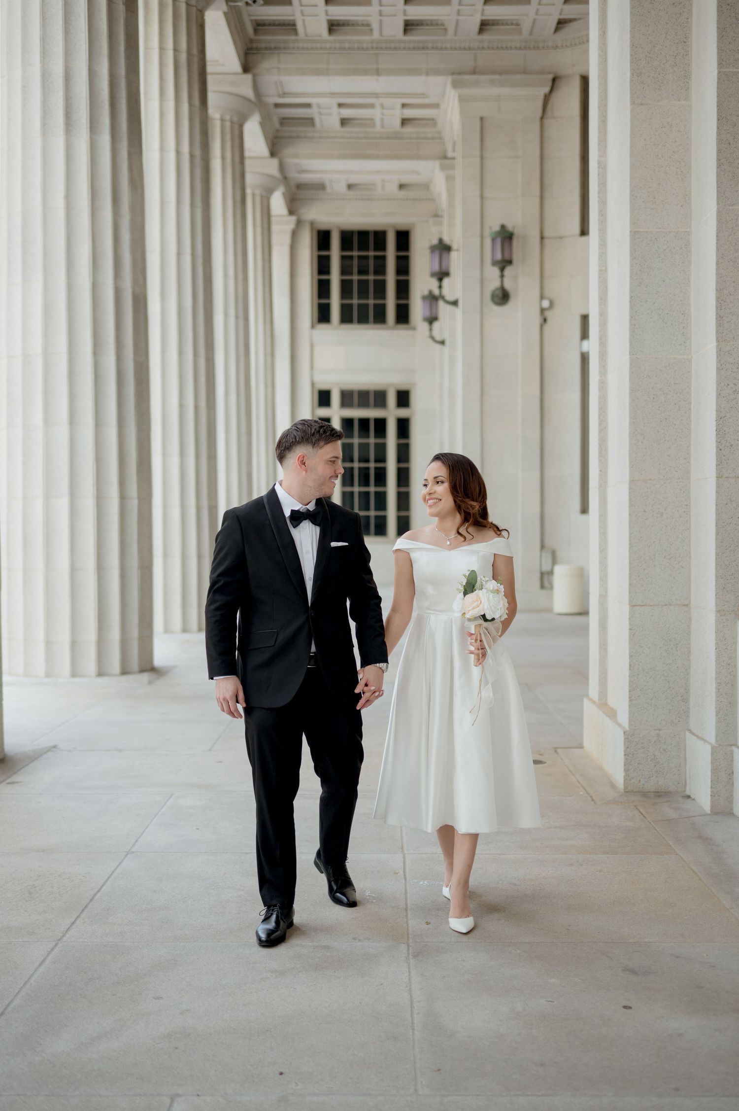 Elegant couple walking through white columned hallway in wedding attire creating a series of romantic poses.