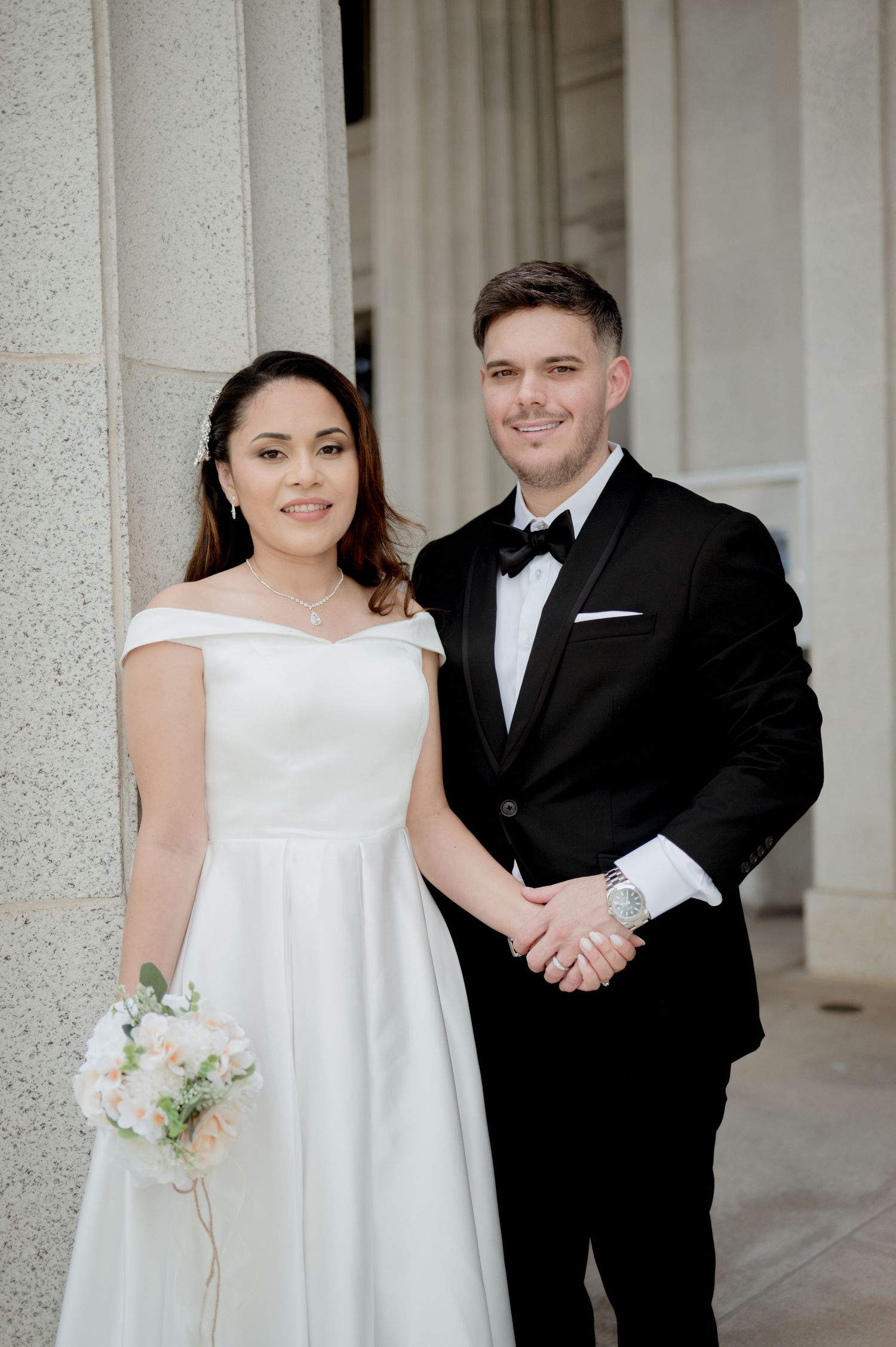 Elegant bride and groom in formal attire posing together in front of classic white stone columns.