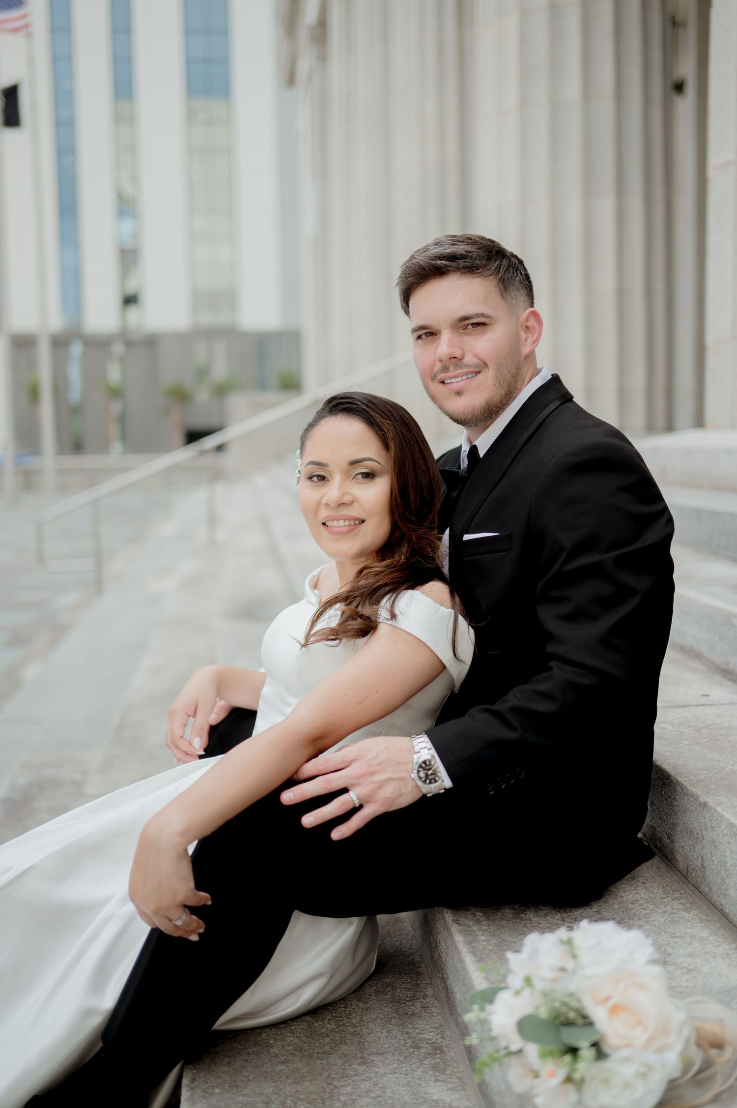 Wedding couple posing together on white marble steps in formal attire with floral bouquet.