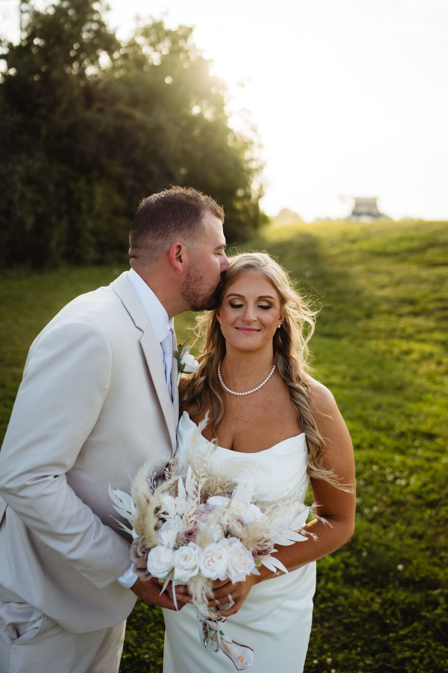 A romantic outdoor wedding photo of newlyweds in white attire with pampas grass bouquet against a sunlit grassy hill.