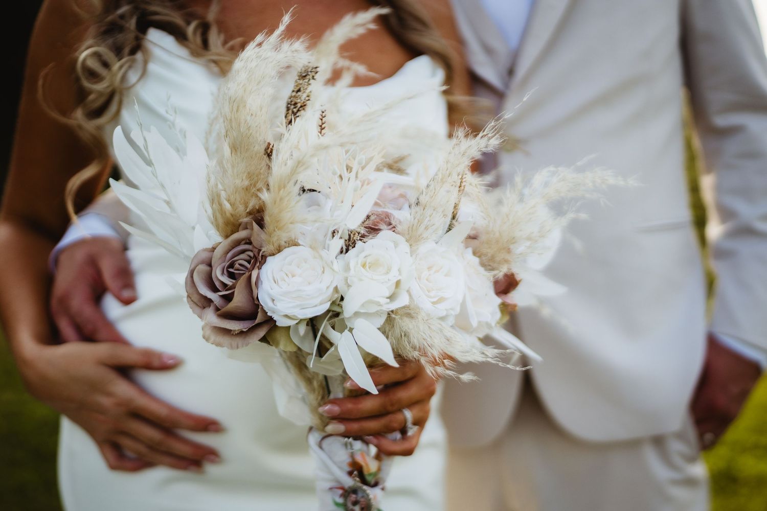 A rustic wedding bouquet featuring white flowers, dried pampas grass and mauve accents held by someone in a white dress.