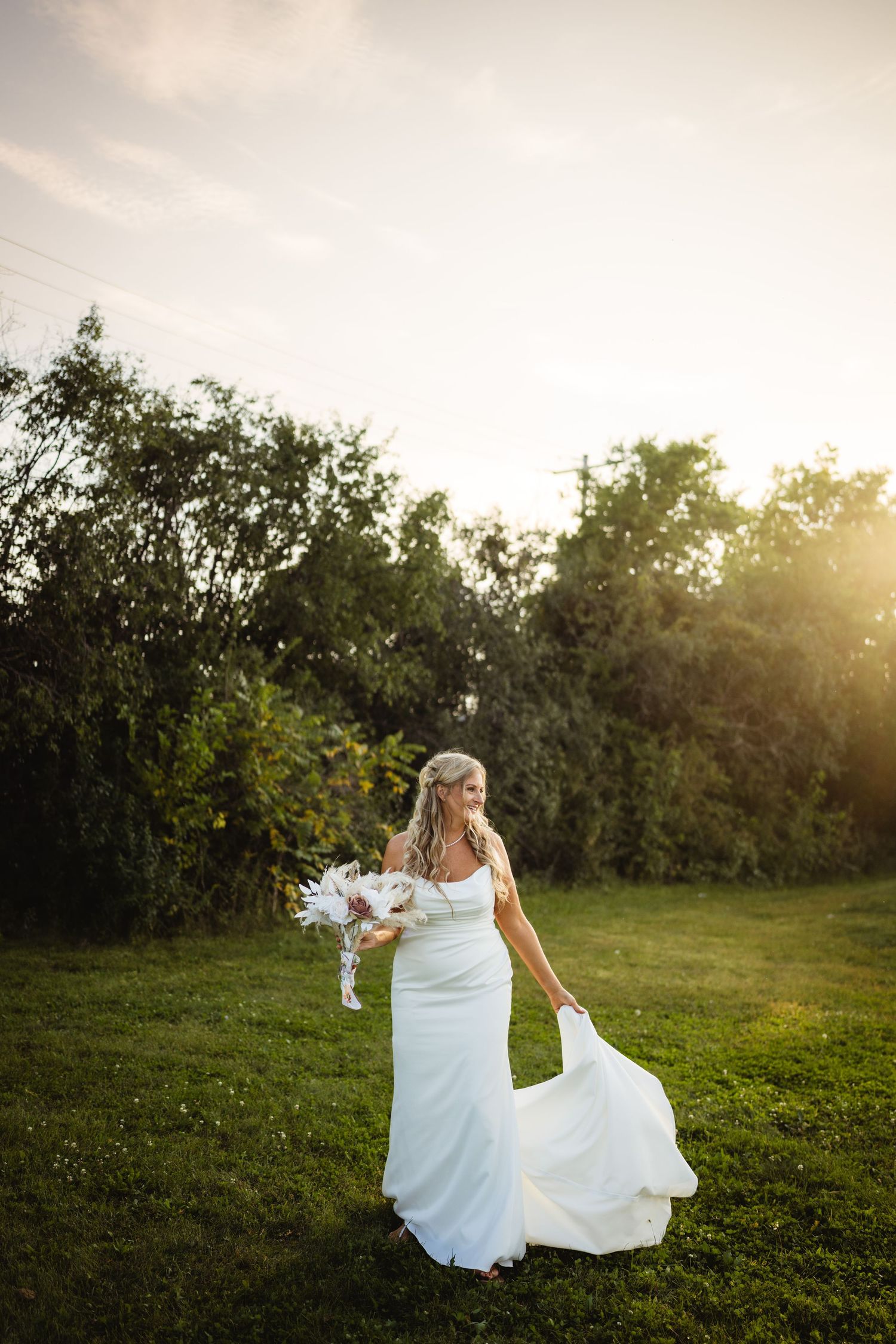 A bride in a white dress twirls joyfully on a grassy hill during golden hour sunset.