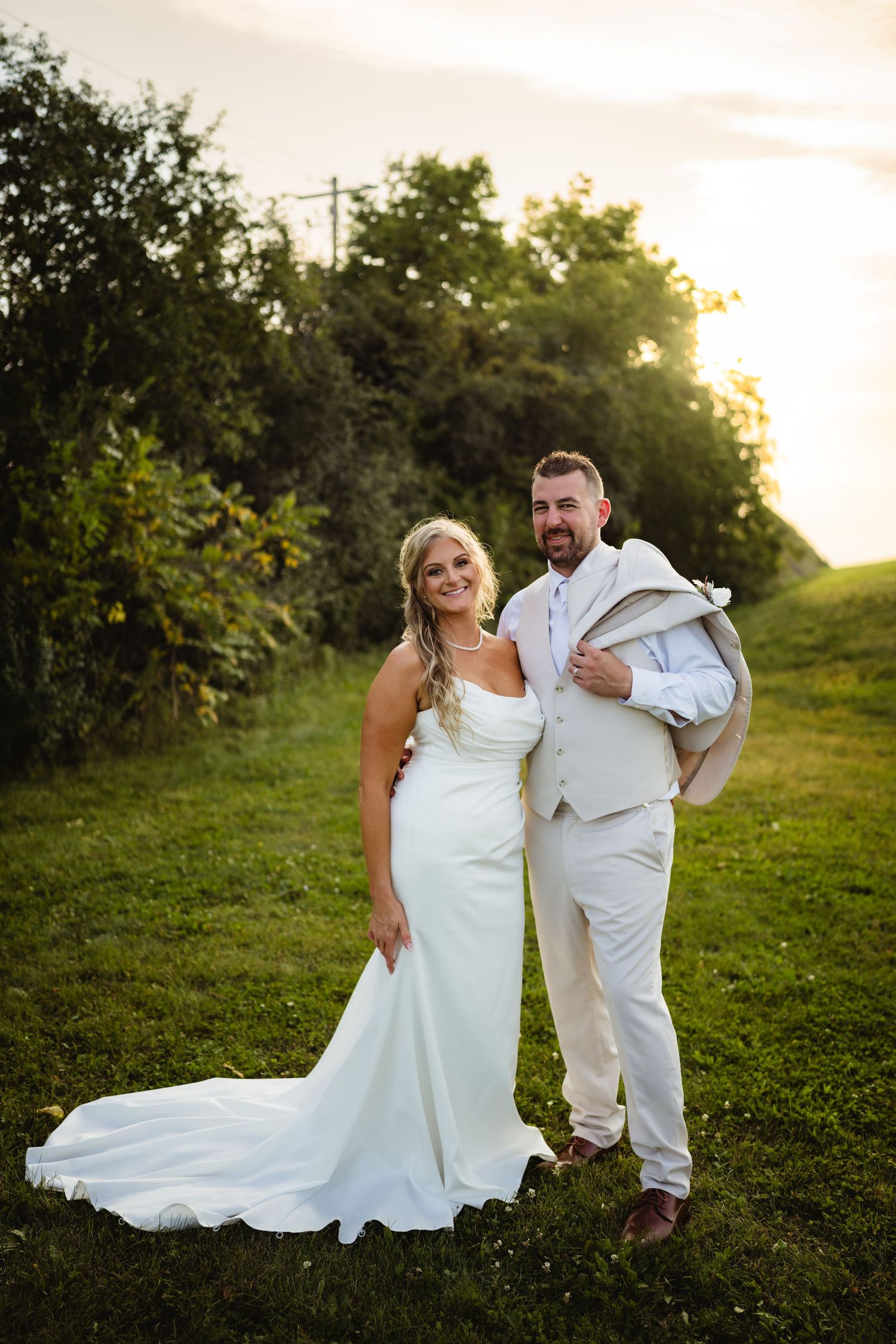 A couple in wedding attire poses together in a lush green field at sunset.