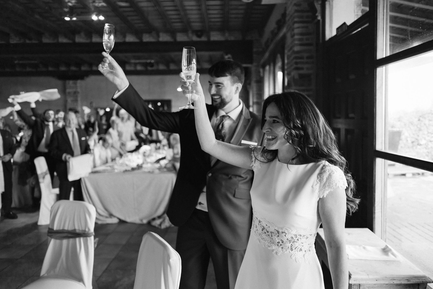 A couple in wedding attire raise their glasses in celebration during a reception party in black and white.