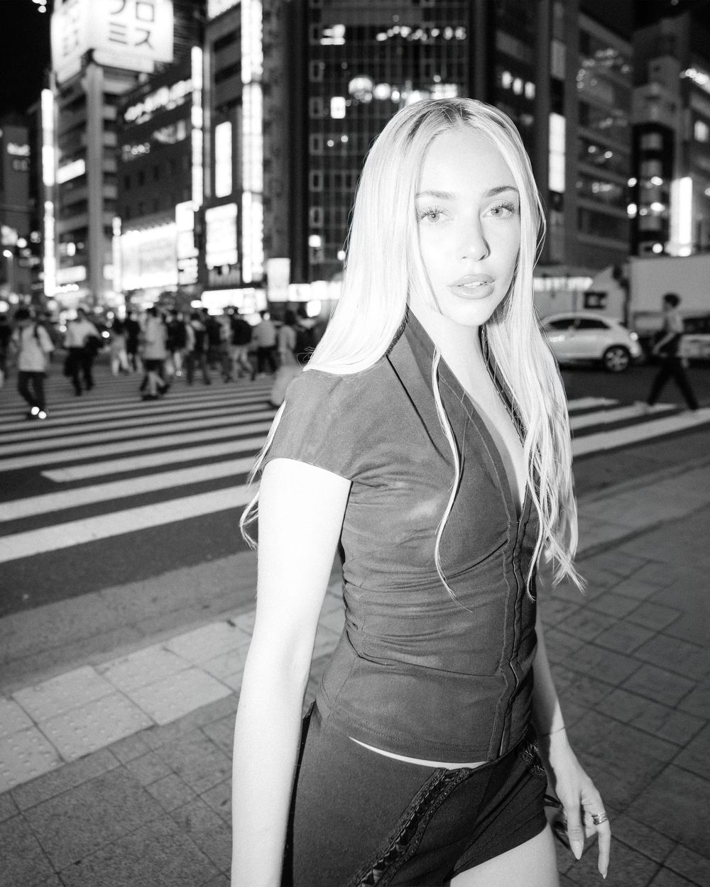 Black and white photo of a blonde woman posing on a busy Tokyo street crossing at night.
