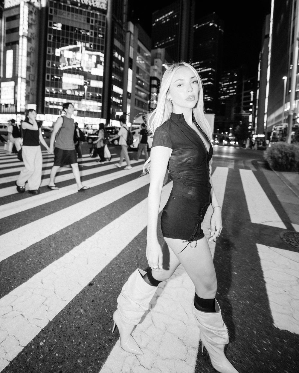 Black and white urban nighttime street photography of a person in a dark outfit posing on a crosswalk in a busy city.