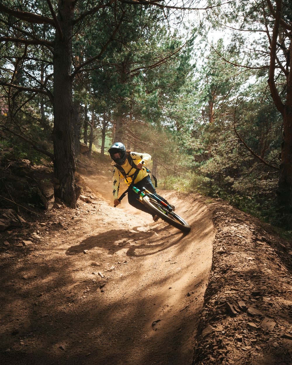 Mountain biker navigates a steep bermed dirt trail through a forest during a downhill run in dynamic sequence.