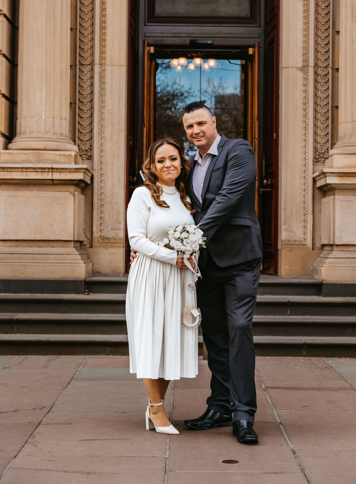 A couple in formal attire shares a playful moment with a corgi dog on stone steps outside an elegant building.