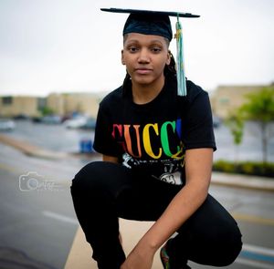 Graduate in black cap and gown poses outdoors with colorful rainbow text on shirt.
