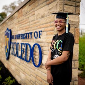 Graduate in cap and gown smiling in front of University of Toledo sign.