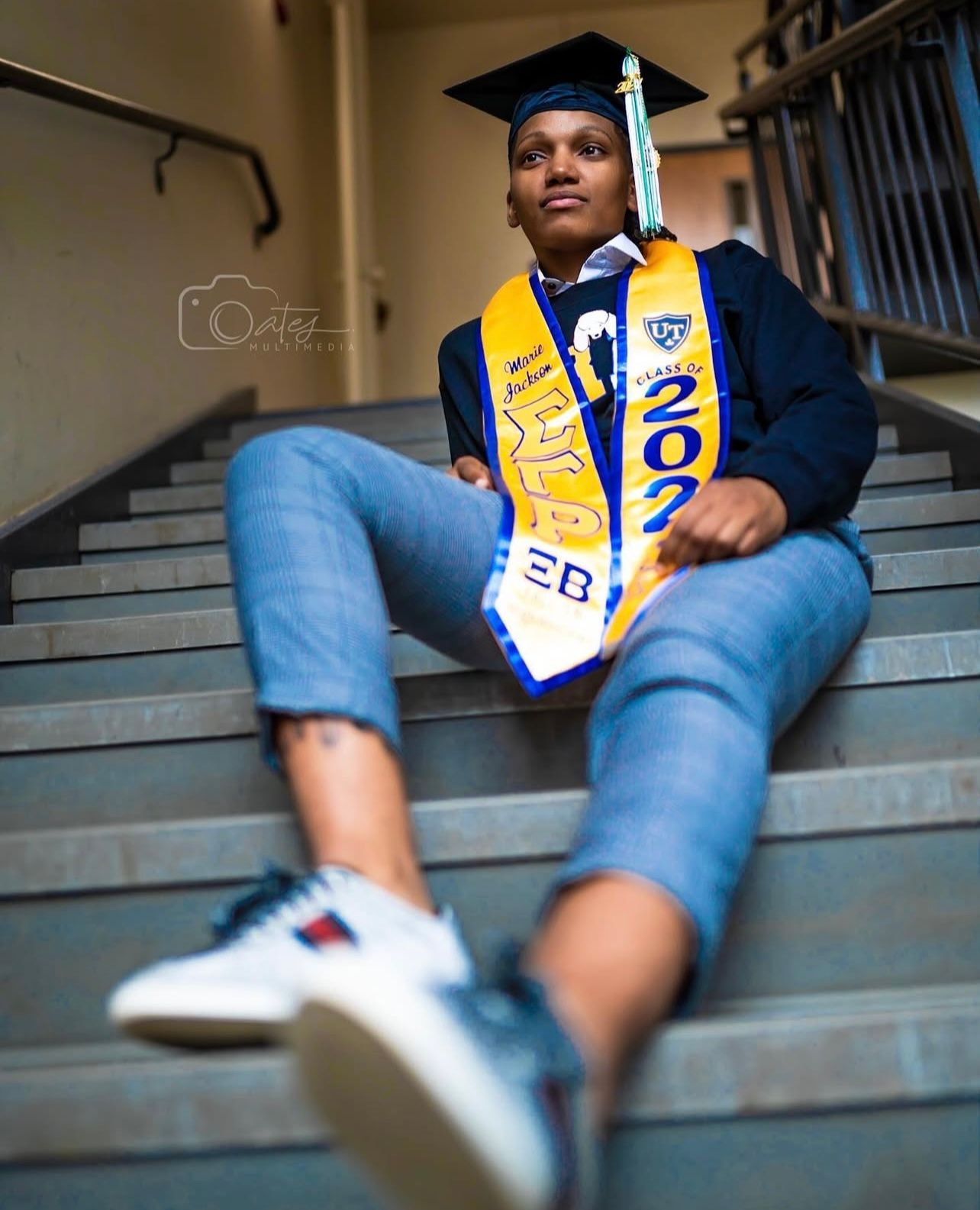 Graduate in cap and gown with class sash sitting on stairs wearing casual jeans and sneakers.