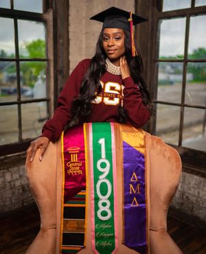 Graduate wearing maroon university sweatshirt with multiple graduation stoles posing in window light.