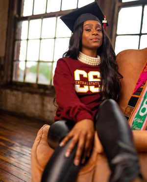 Graduate wearing maroon jersey and pearl necklace poses for professional graduation photos in rustic setting.
