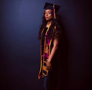 Dramatic studio portrait of graduate in cap and gown with honor stoles against dark background.
