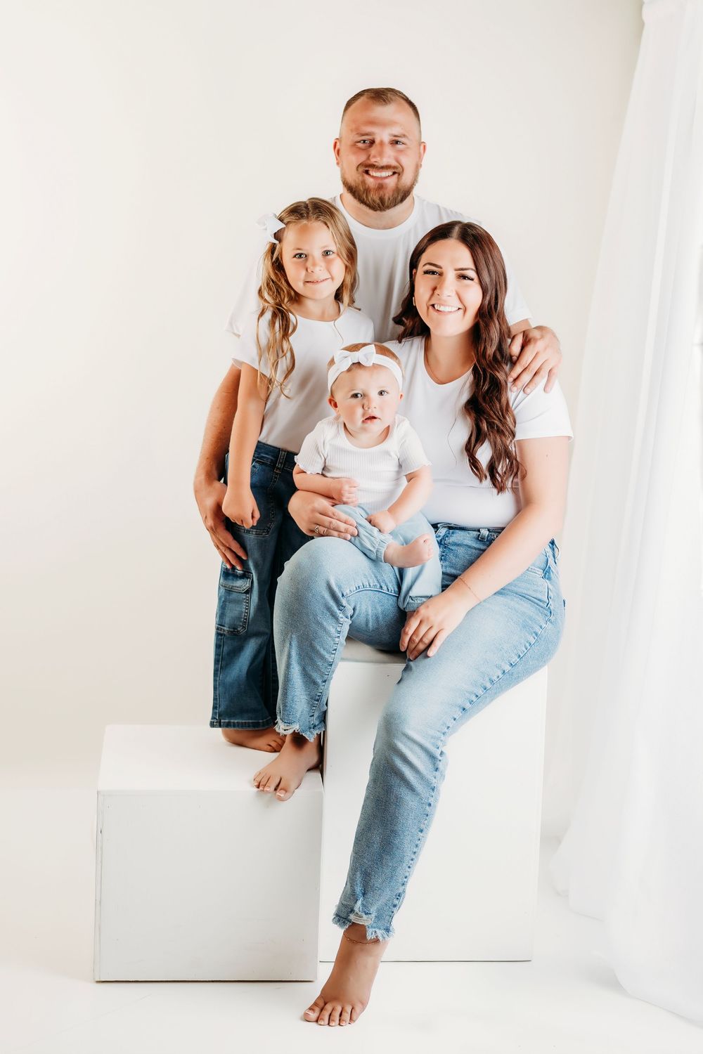 A family wearing casual denim and white shirts sits together on a white block in a minimalist studio setting.