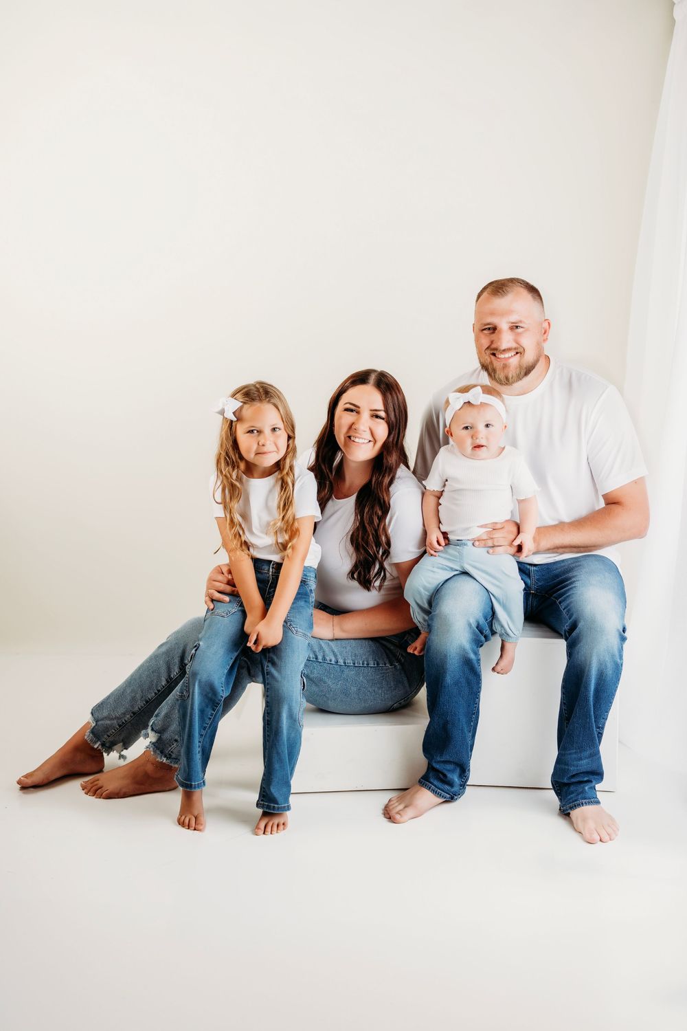 A family in coordinated casual denim and white outfits sits together on a white chair in a bright studio setting.