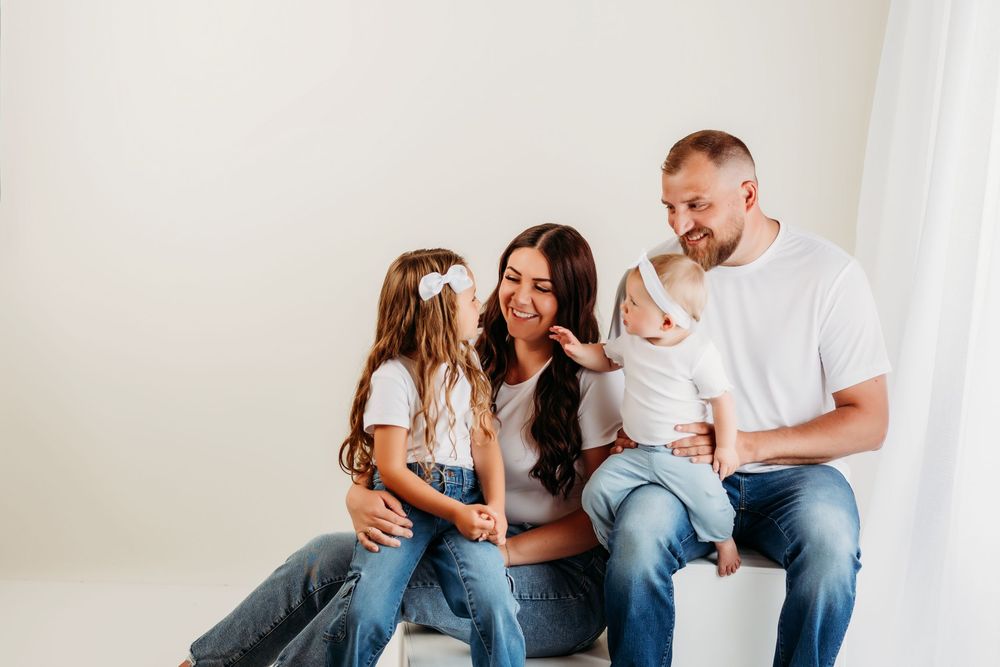 Happy family sits together on floor against white wall wearing casual outfits of jeans and t-shirts.