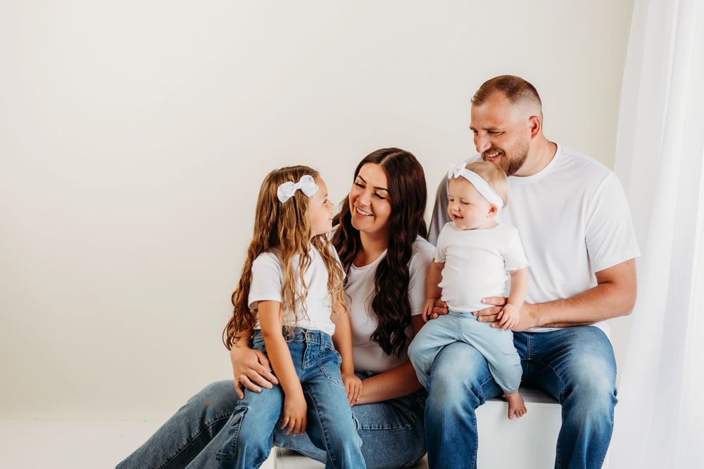 Family of four poses casually together against bright white wall in matching denim and white outfits.