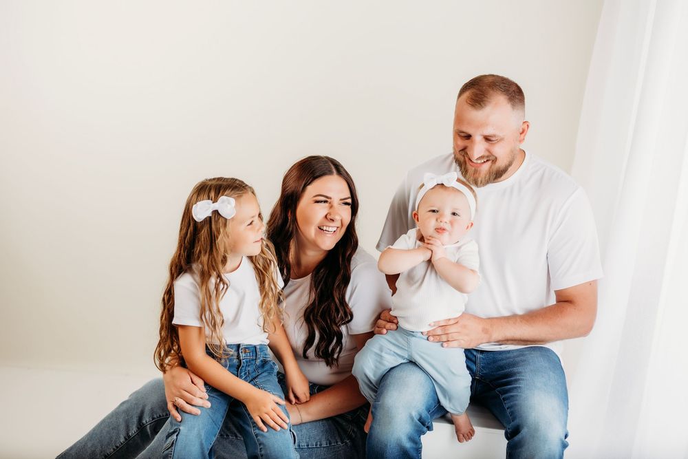 A happy family wearing white and blue outfits sits together on a light-colored couch for casual photos.