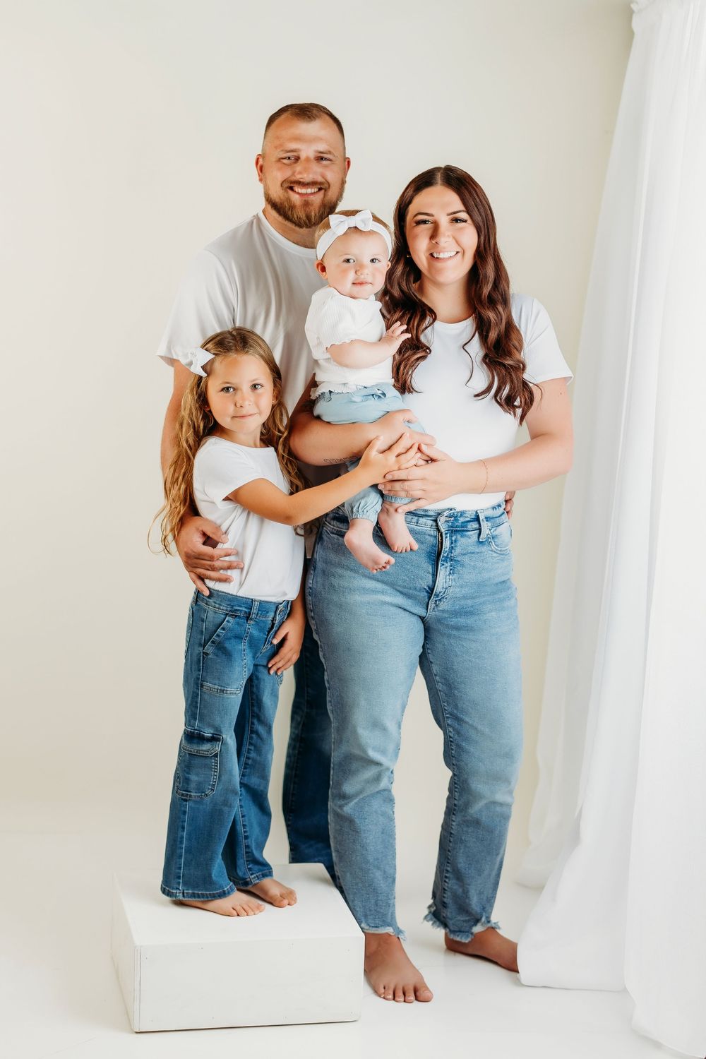 Family wearing matching white shirts and blue jeans poses together against white backdrop in studio setting.