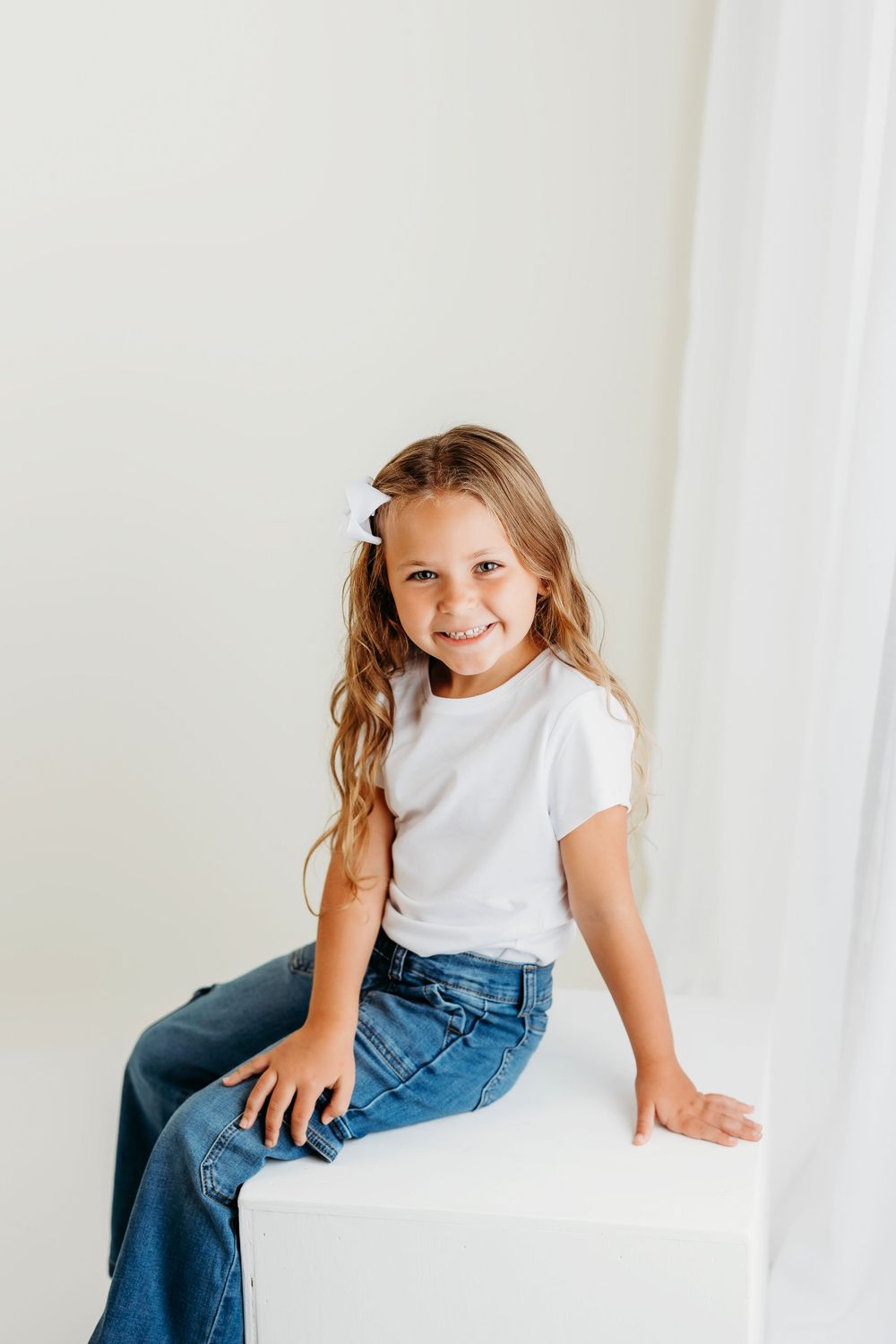 Young child wearing white t-shirt and blue jeans sitting on white surface against light background.