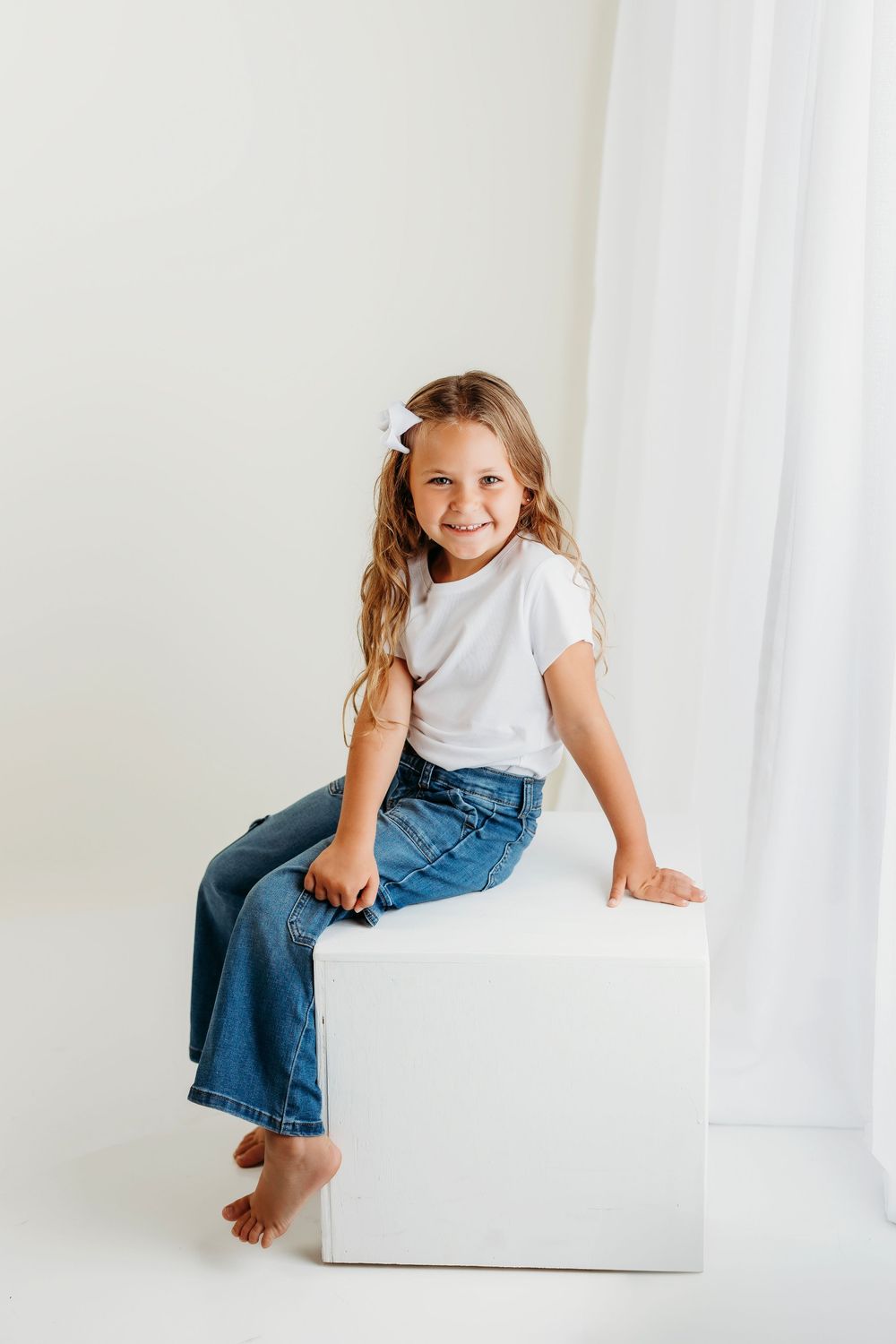 A child in blue jeans and white t-shirt sits casually on a white block against a light studio background.