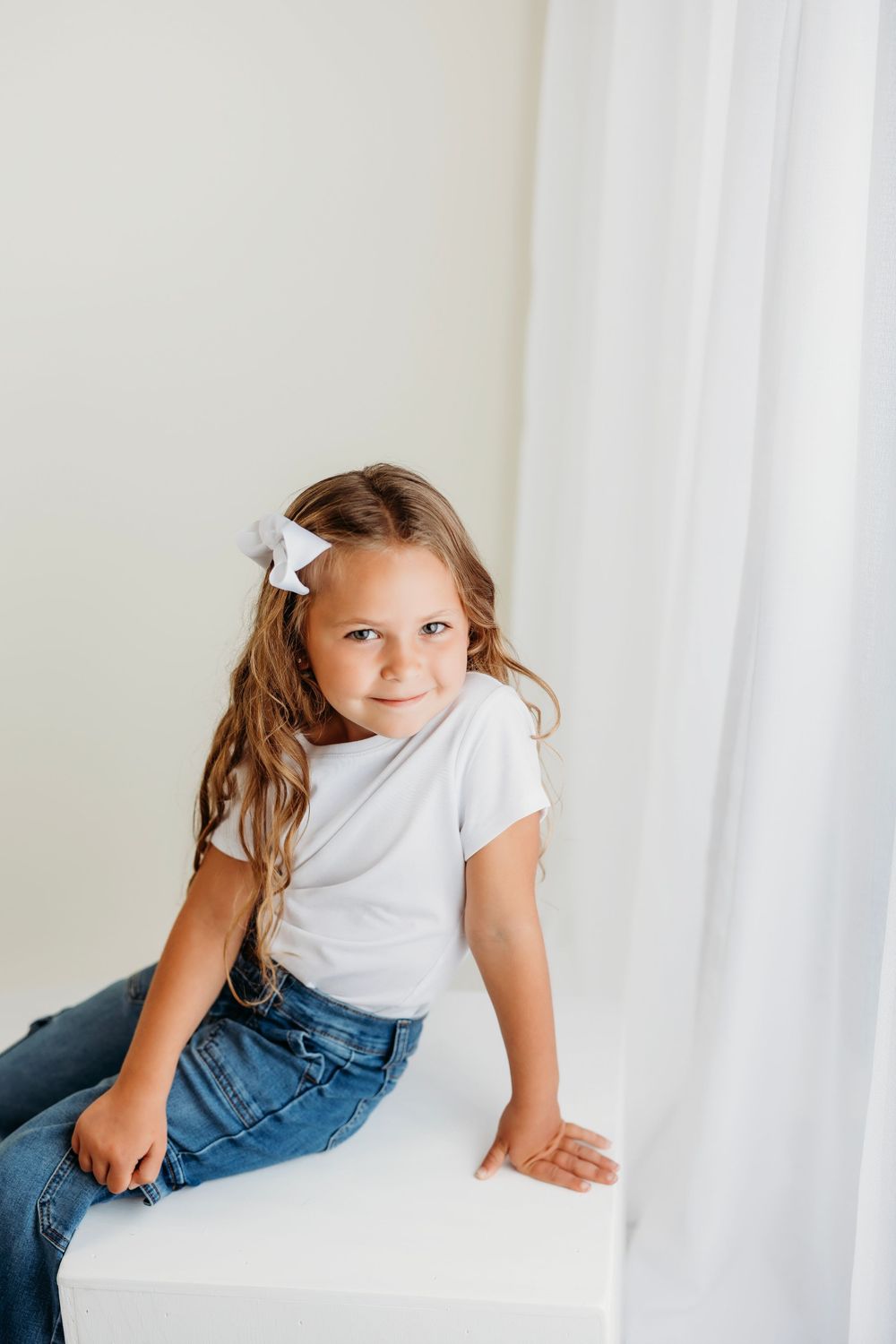 Young child with brown hair wearing white shirt and jeans lounges against white backdrop in natural light setting.