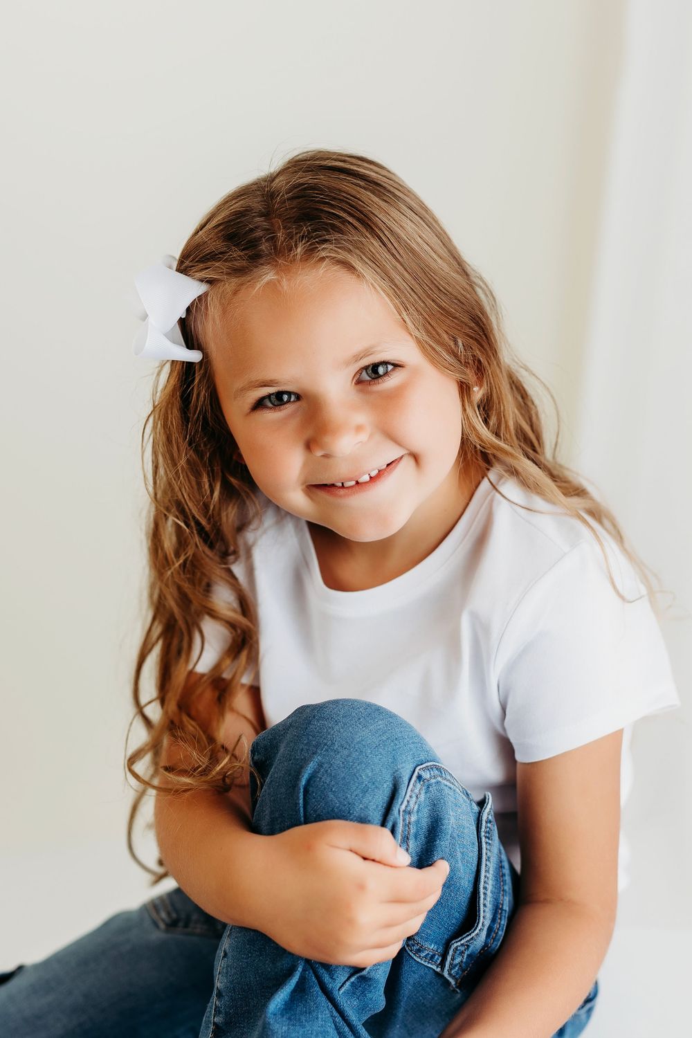 A child wearing a white t-shirt and jeans sits on a bed while looking at the camera with a bright smile.