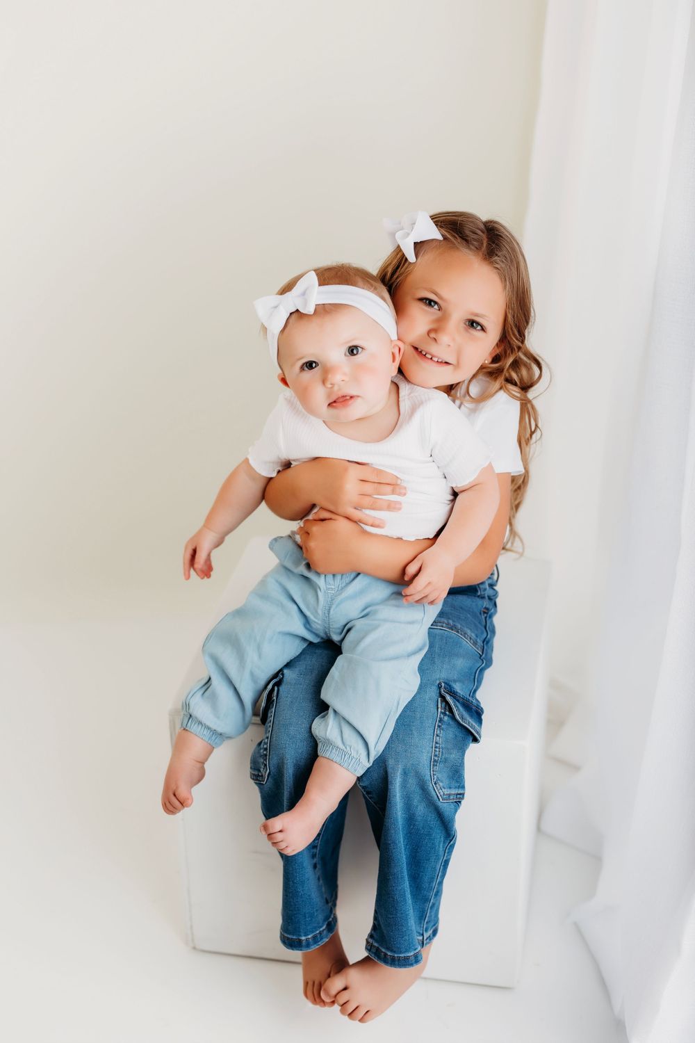 An infant in a white top and blue jeans sits in a bright white studio space.