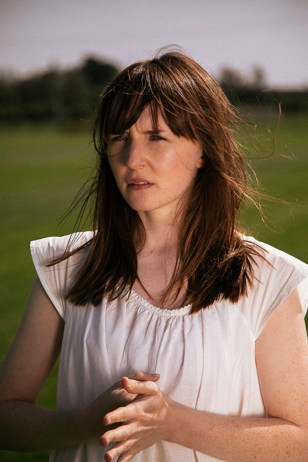 Young woman with brown hair in white sleeveless top standing outdoors in a green field on a windy day.