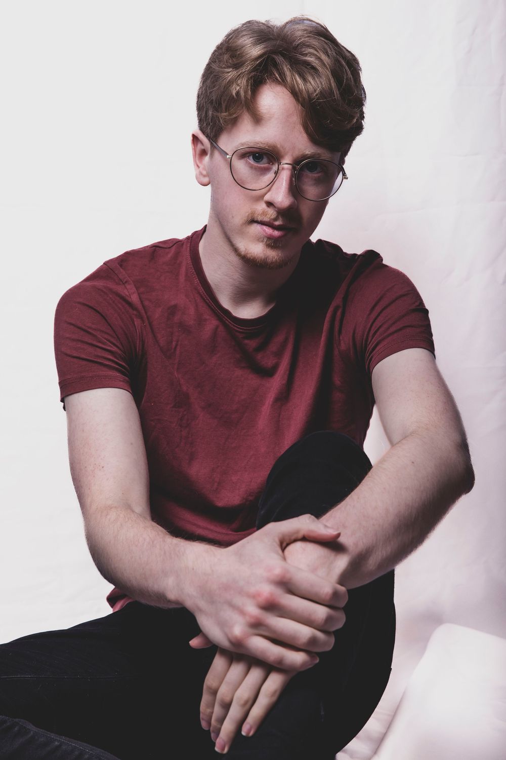 Young man with glasses and red t-shirt sitting relaxed against white background in casual portrait photo.
