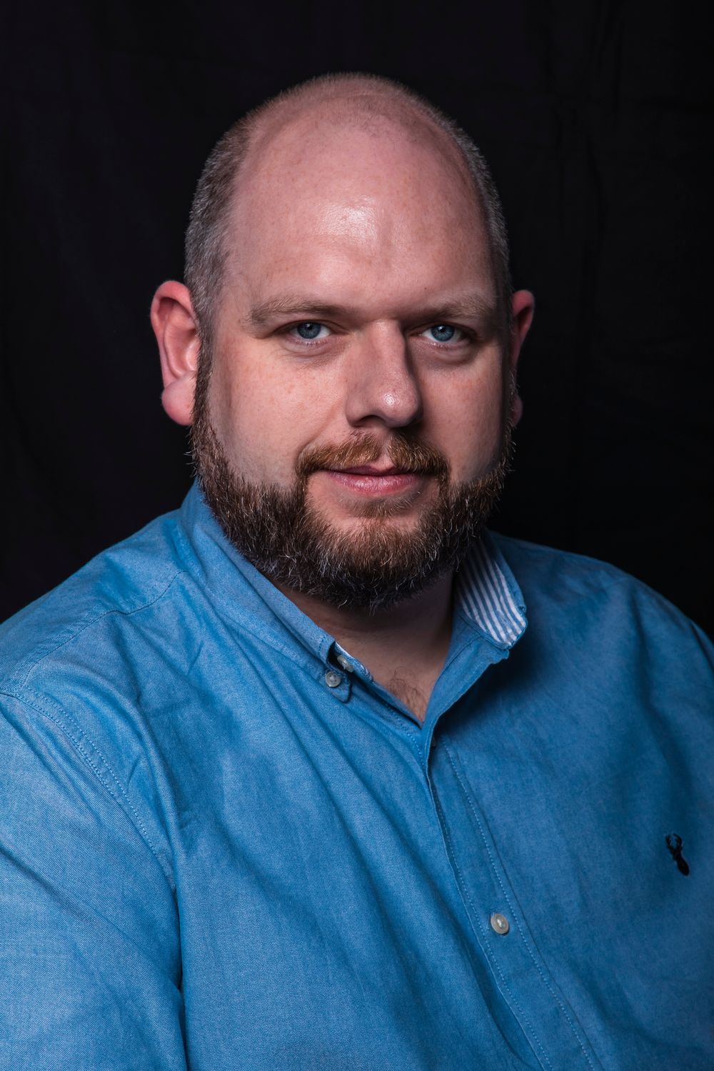 Bald bearded man in blue chambray shirt poses for professional portrait against dark background.