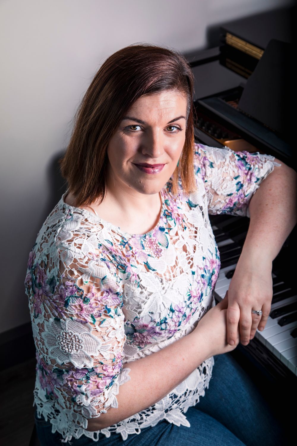 Woman in floral lace top smiling beside a grand piano in a bright studio setting.