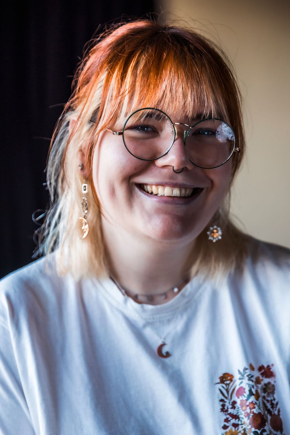 Smiling young woman with red hair, round glasses, floral earrings, and a white embroidered t-shirt.