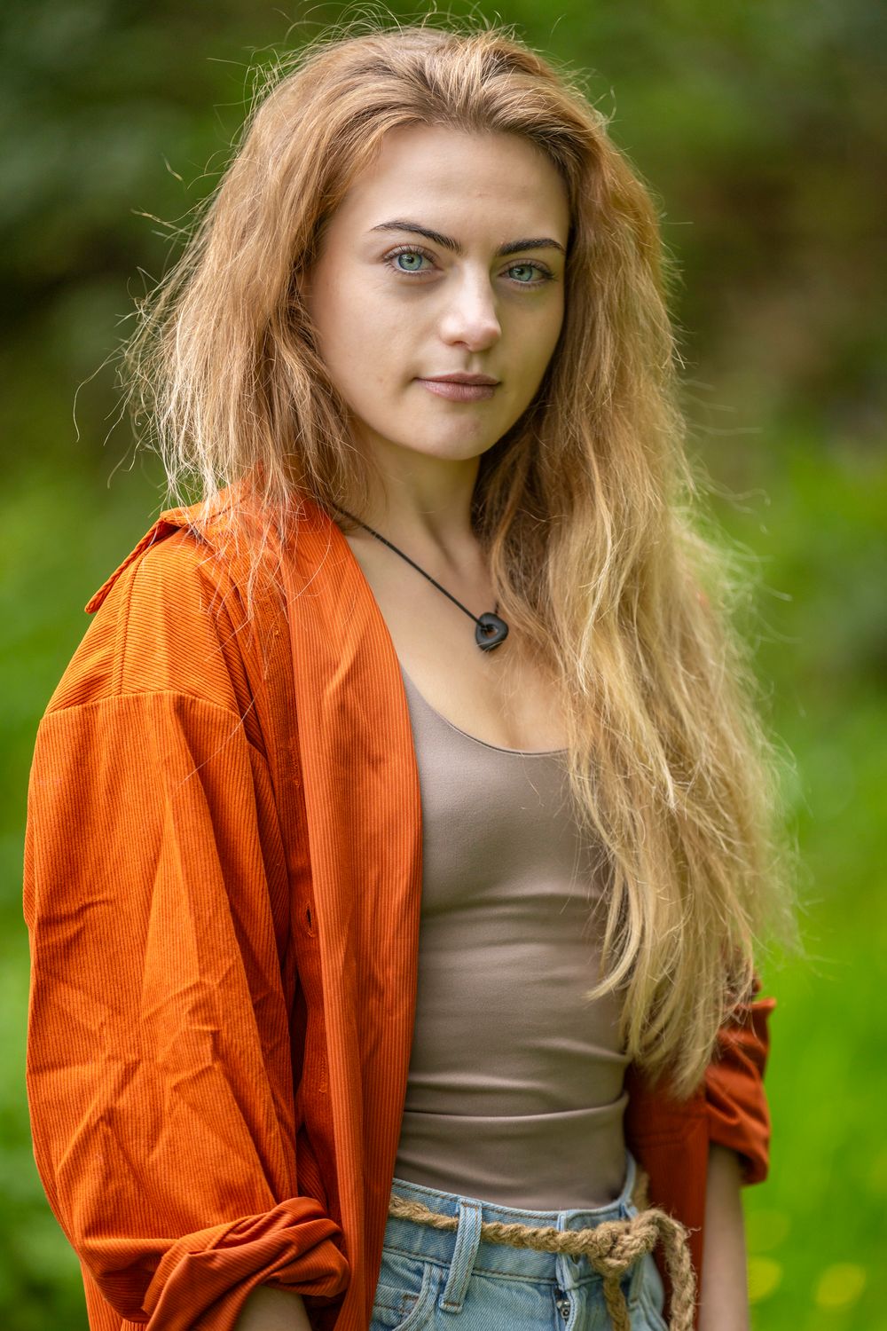 Young woman with long wavy auburn hair and blue eyes wearing an orange shirt and beige tank top outdoors.