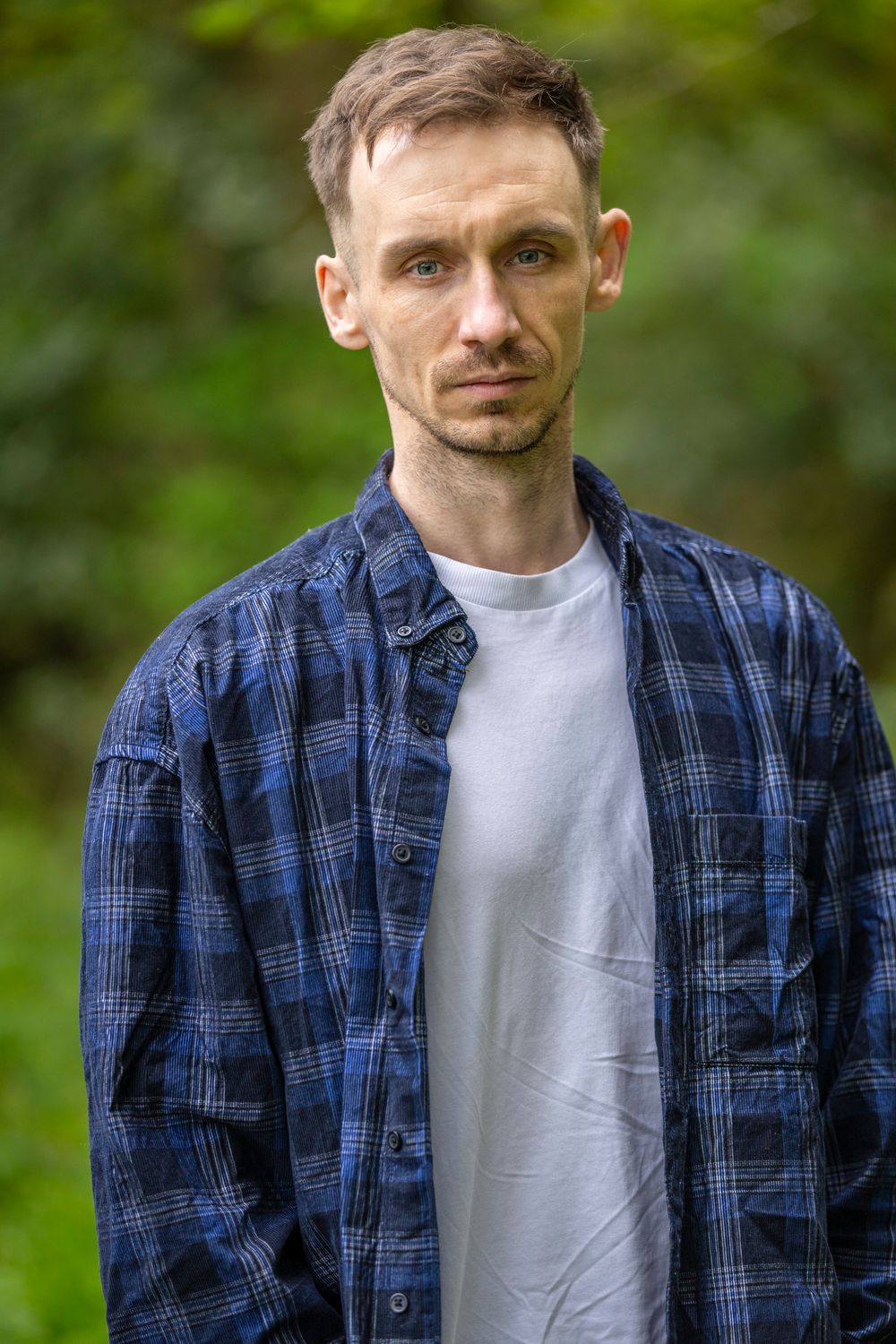 A young man in a blue plaid shirt over a white tee stands outdoors with green foliage in the background.