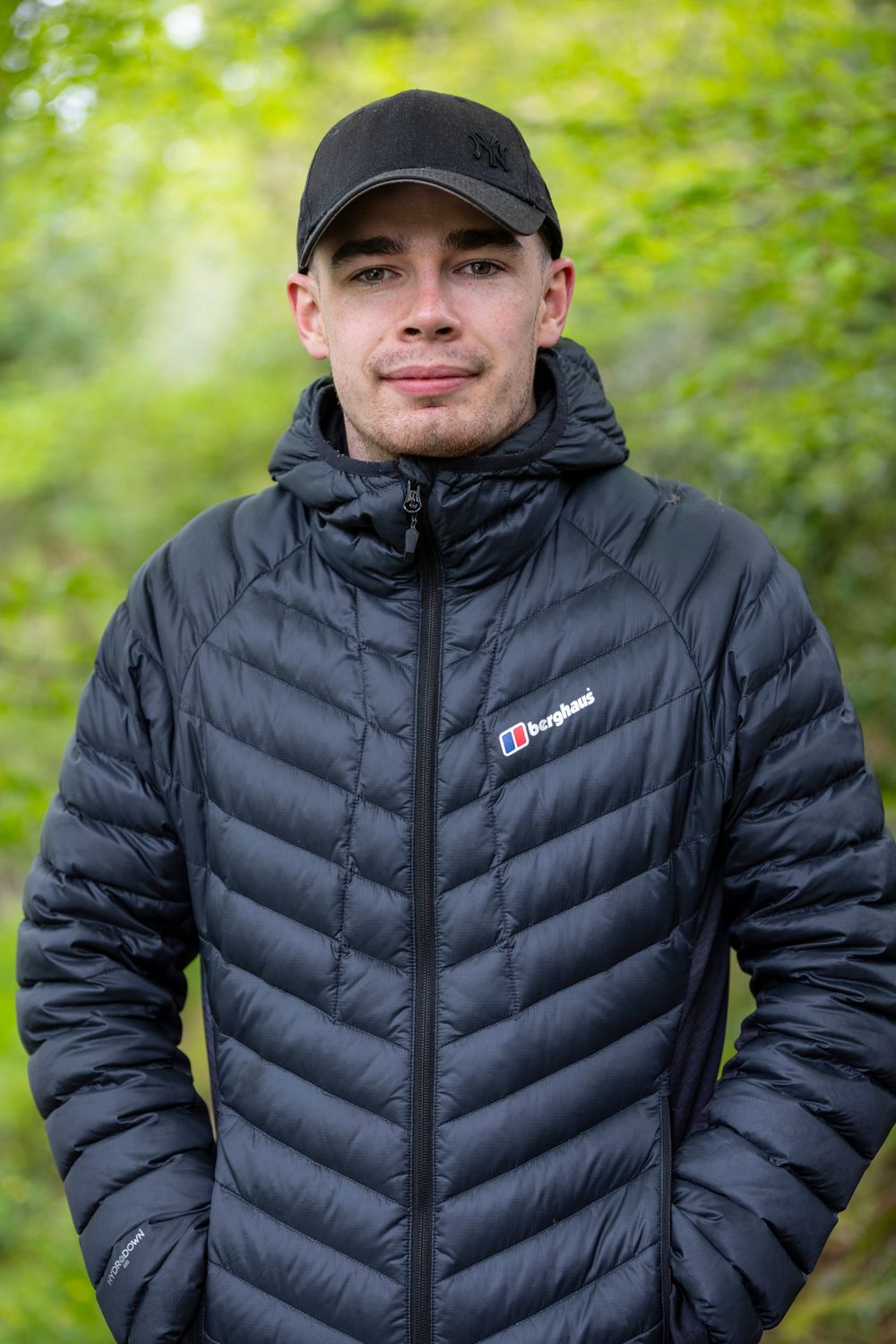 Young man wearing a black Berghaus puffer jacket and cap, standing outdoors in a green woodland setting.