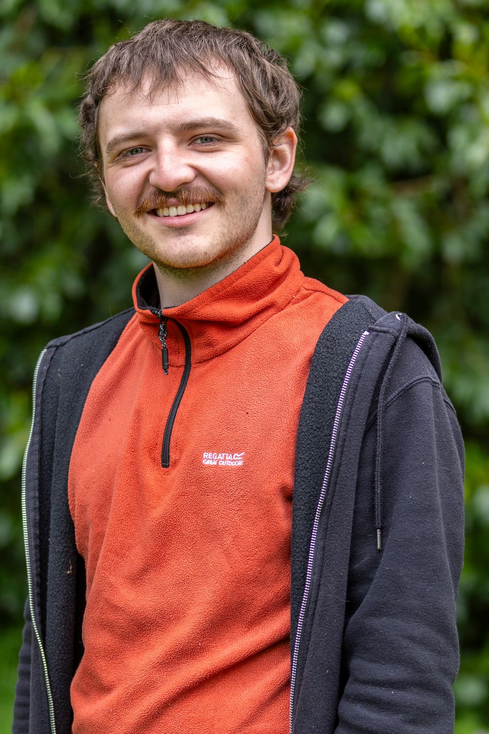 Smiling young man with stubble wearing a red Regatta fleece vest and black jacket, standing outdoors near green foliage.
