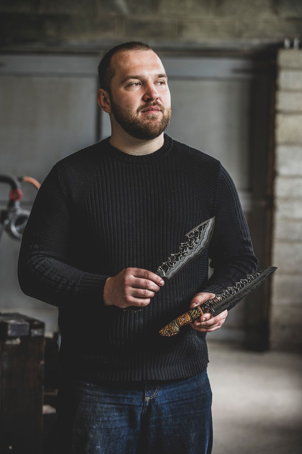 Bearded man in black sweater holding an ornate decorative knife in a rustic workshop setting.