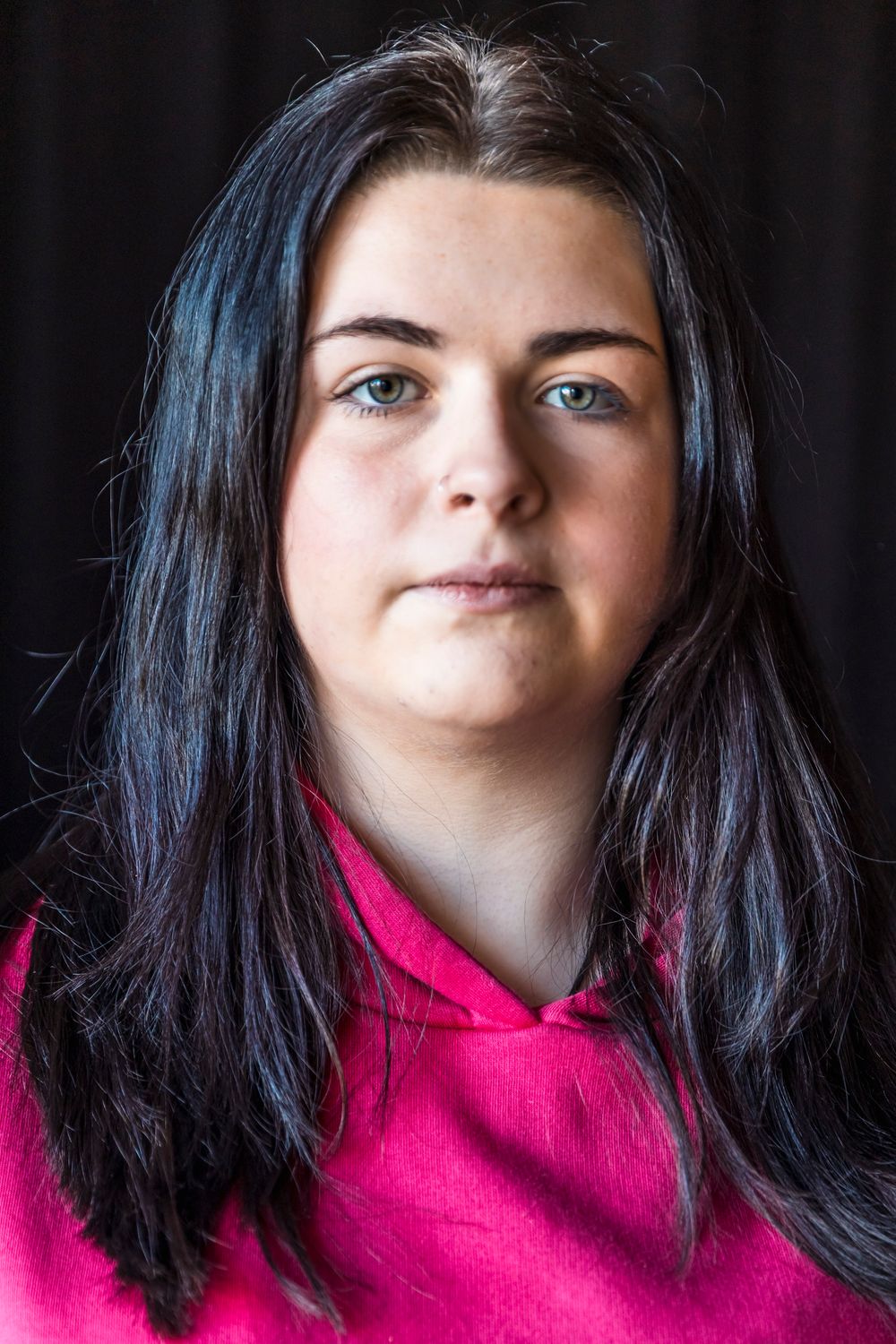 Young woman with dark hair and blue eyes wearing a pink hoodie, posed against a dark background.