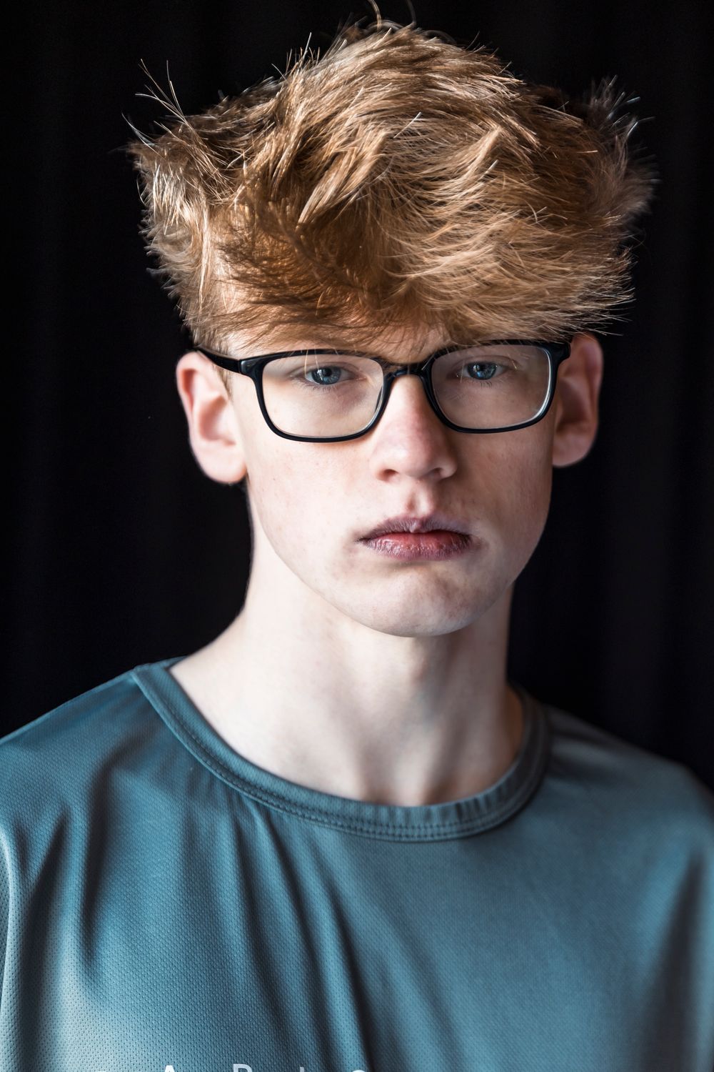 Young redhead teen boy with messy hair wearing black rectangular glasses and gray t-shirt against dark background.