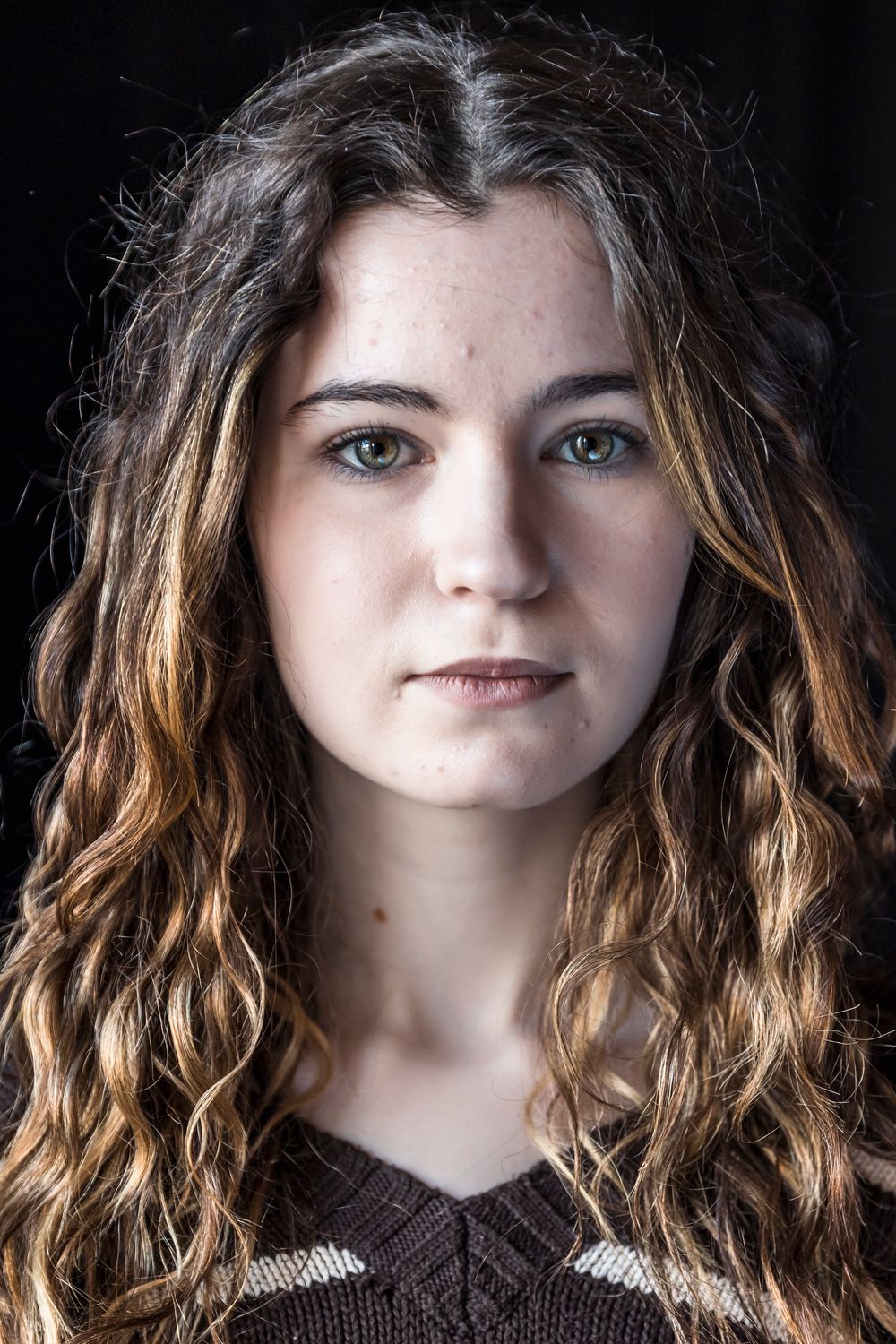 Young woman with long curly brown highlighted hair and green eyes posing against a dark background.