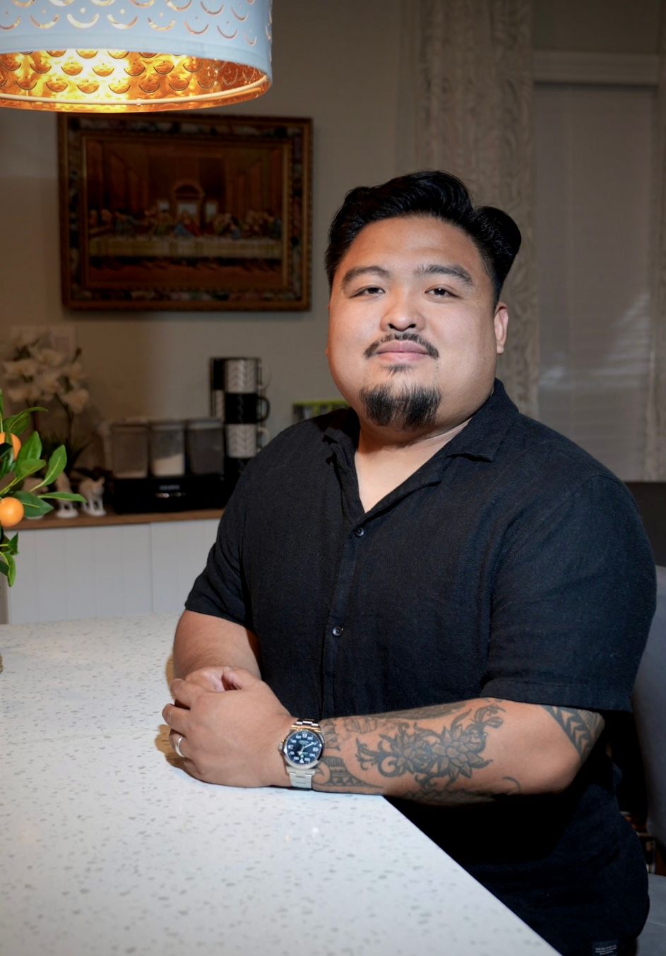 Portrait of a man in a black shirt sitting at a kitchen counter with a confident smile.