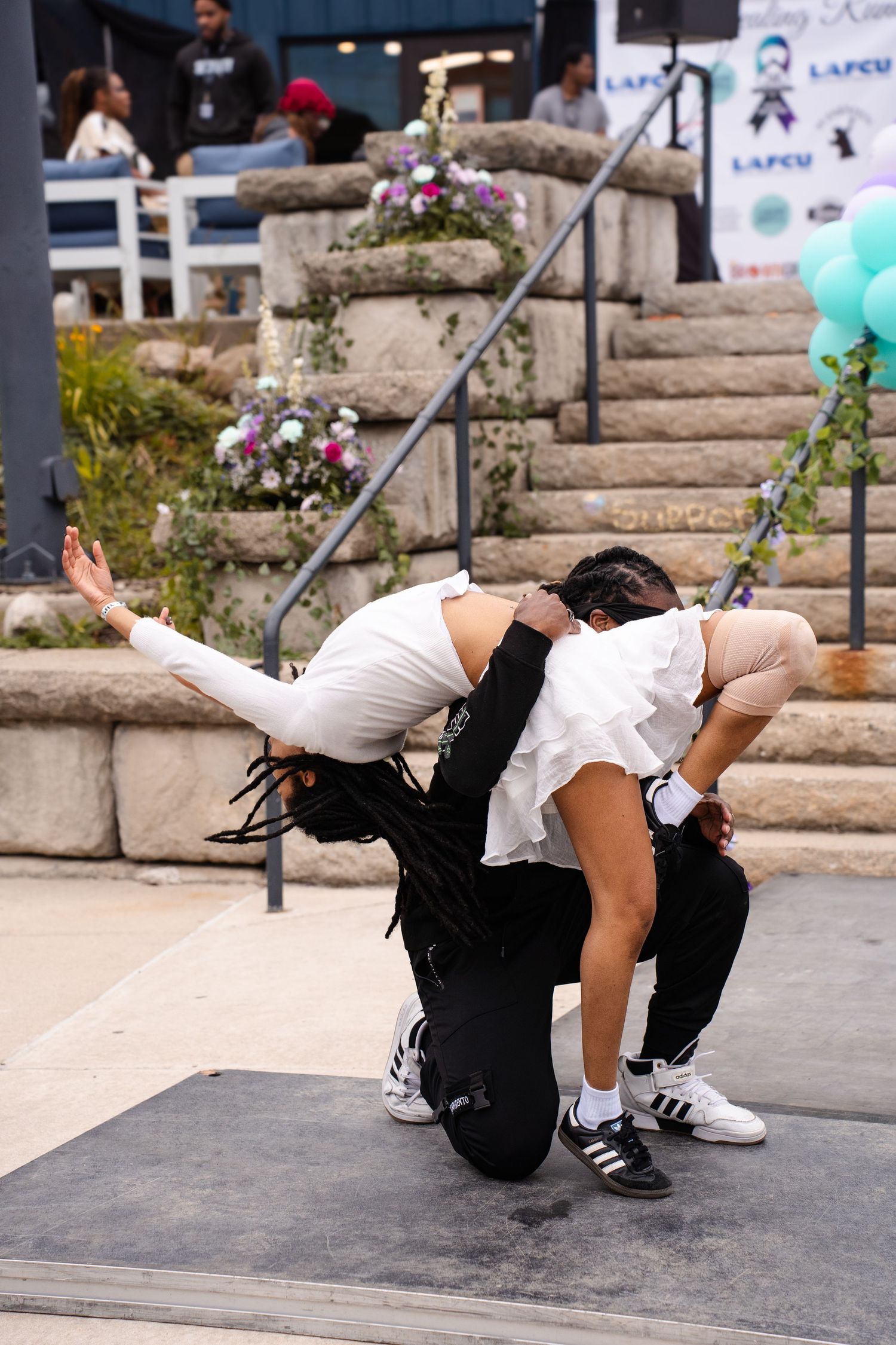 Dancer in white top and black pants performs dynamic breakdance moves on outdoor concrete stage with stone steps.