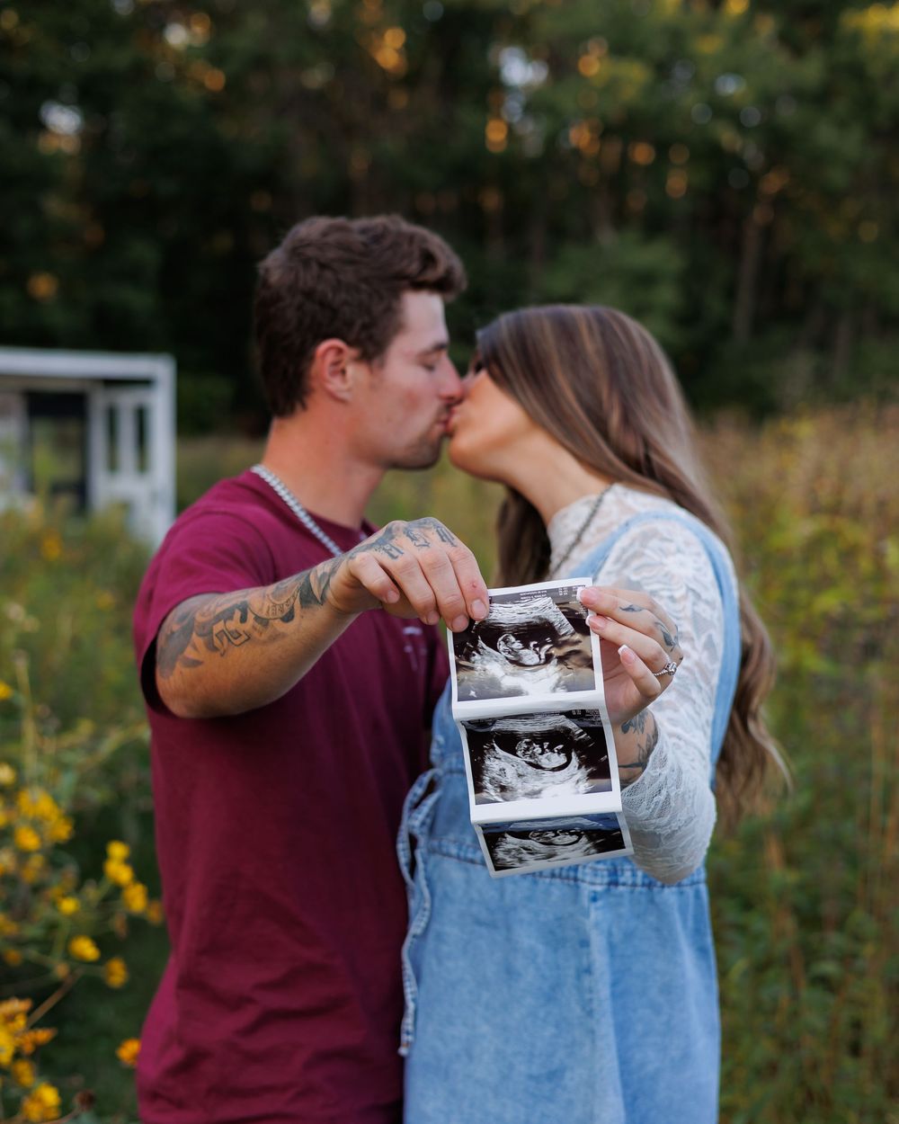 A couple sharing a kiss while holding up an ultrasound photo in an outdoor setting with wildflowers and greenery.