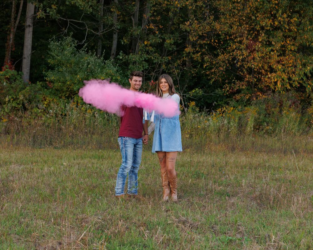 A couple stands in a grassy field with pink smoke floating in the autumn air during a special moment.