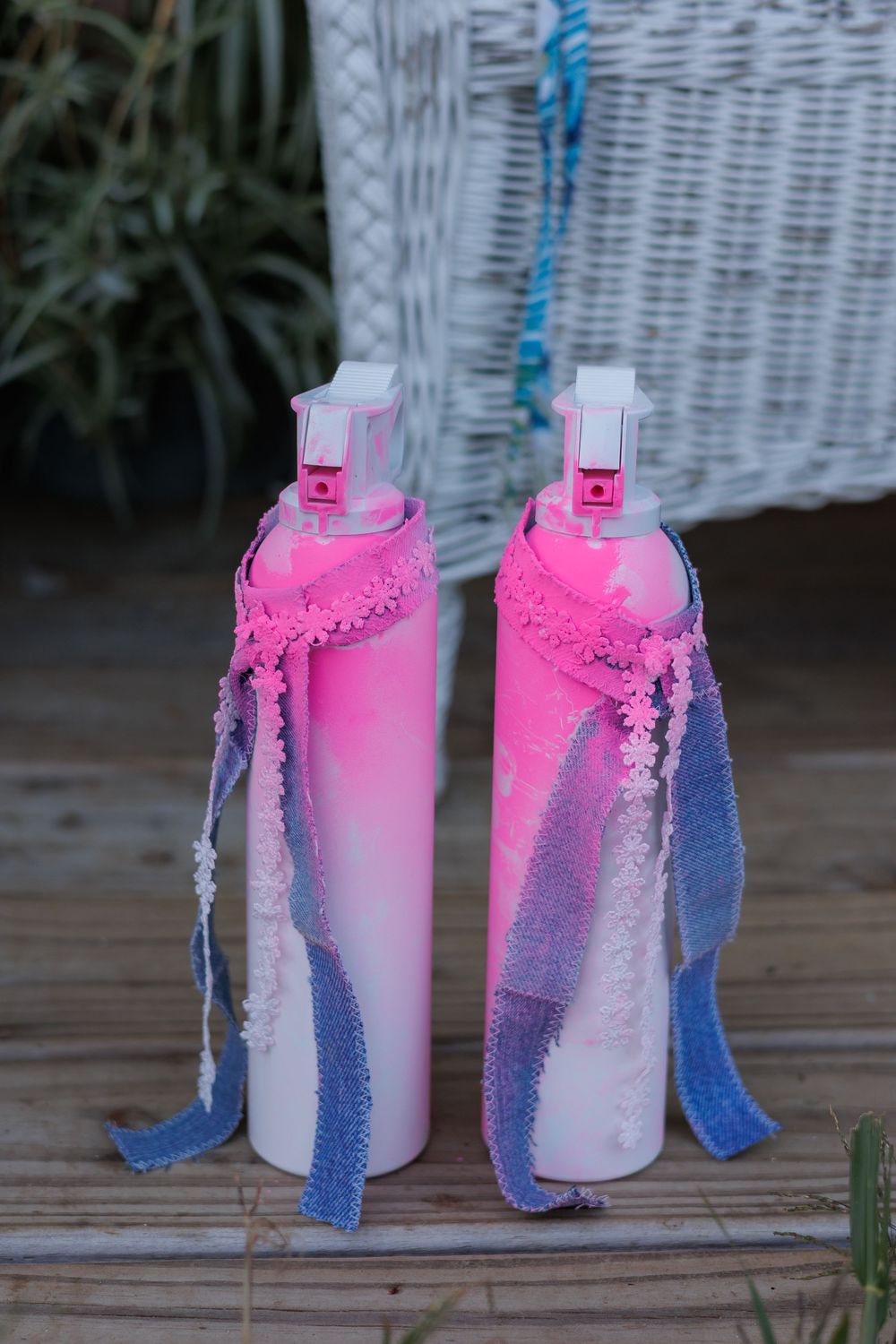Pink and blue tie-dye spray bottles with decorative lace trim sit on a wooden surface against a white wicker background.