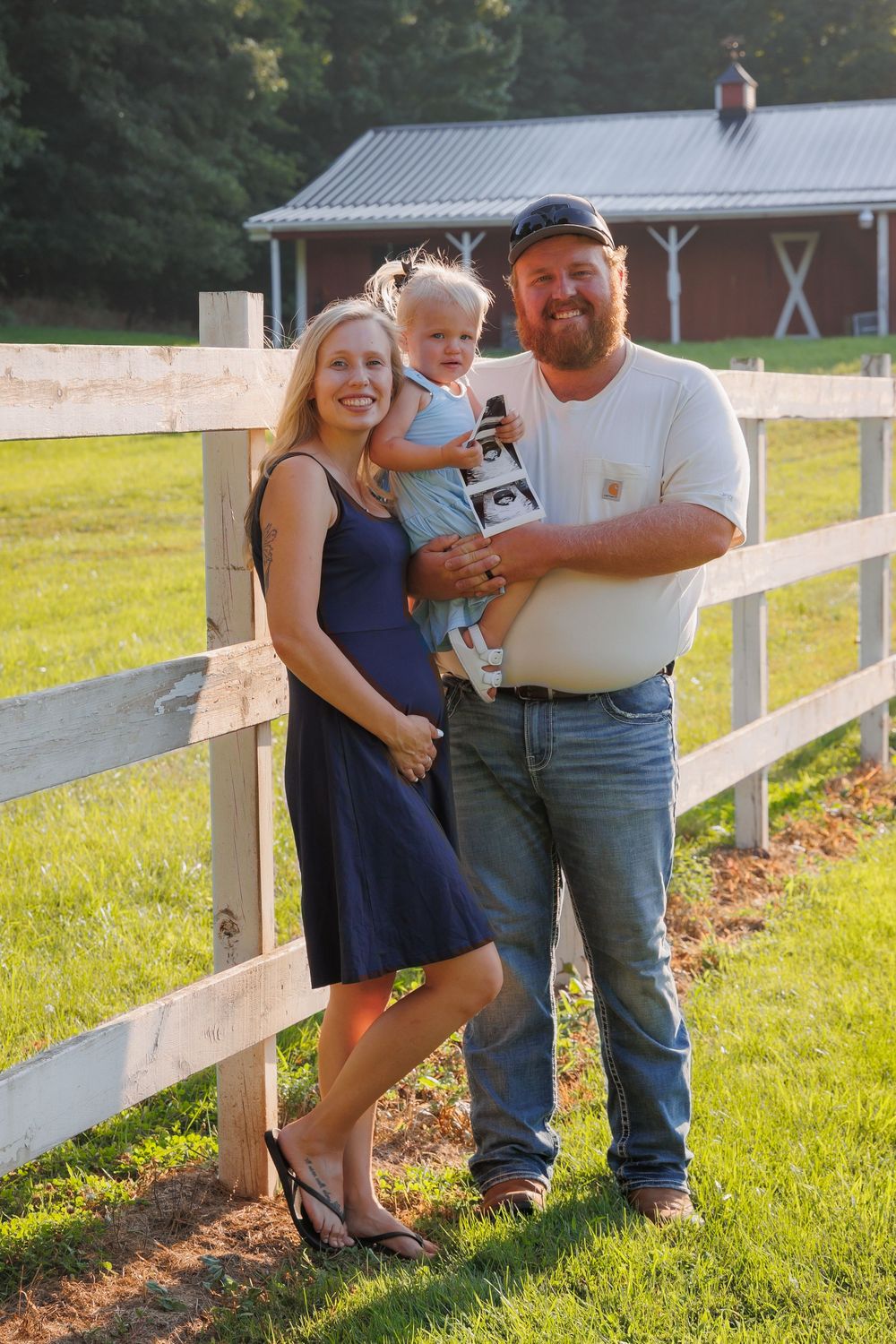 A family stands together by a white fence on a sunny day with a red barn building in the background.