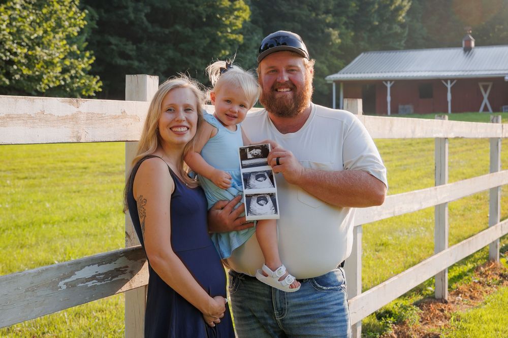 A happy couple in casual attire stands outdoors by a white fence while holding an ultrasound photo together.